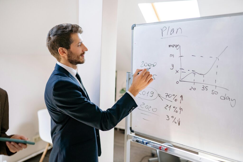 A businessman in a suit writes financial data on a whiteboard during an office planning session.